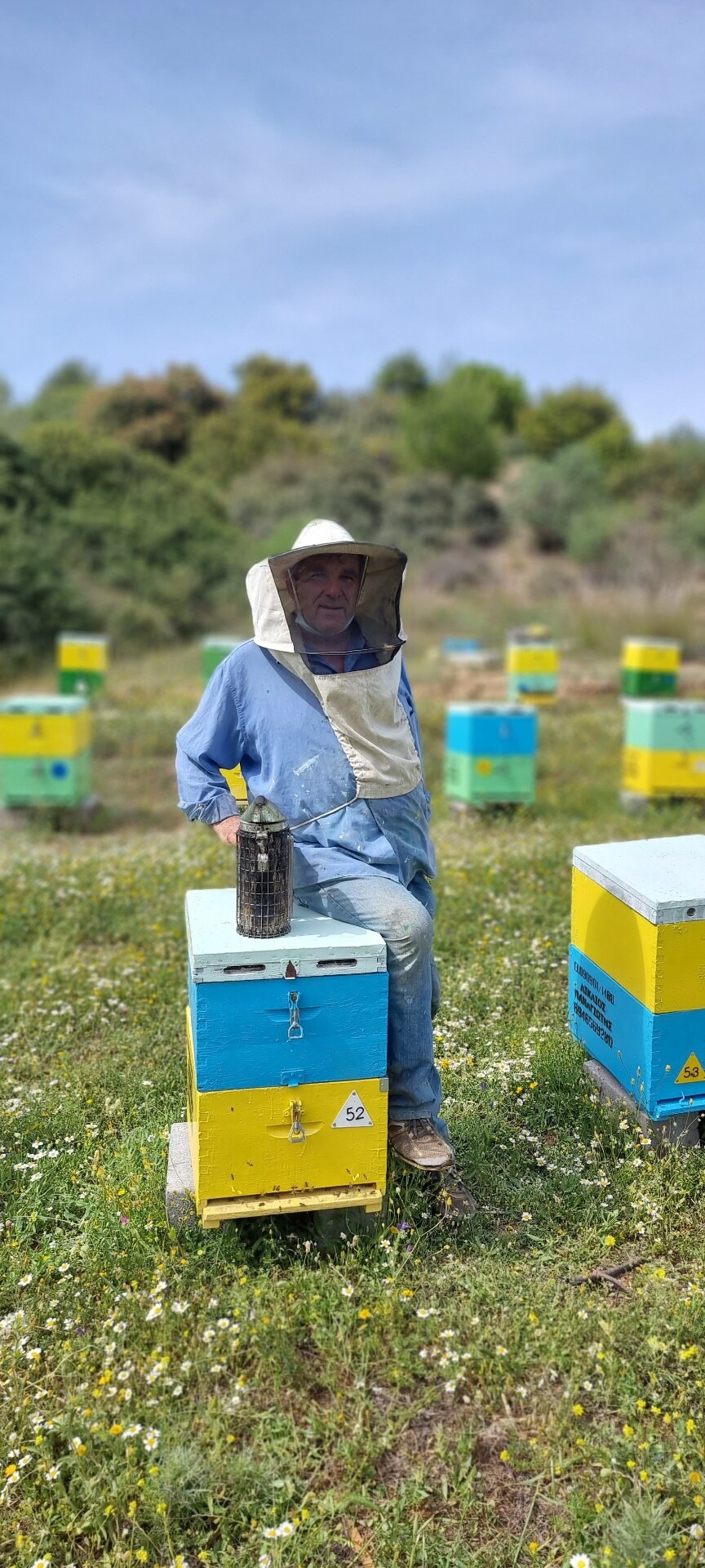 A beekeeper seated on a hive in a flower-filled meadow.
